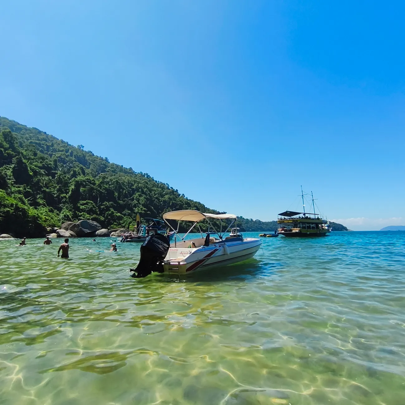 Lancha Oceania fundeada em água turquesa transparente, com ilha e escuna ao fundo